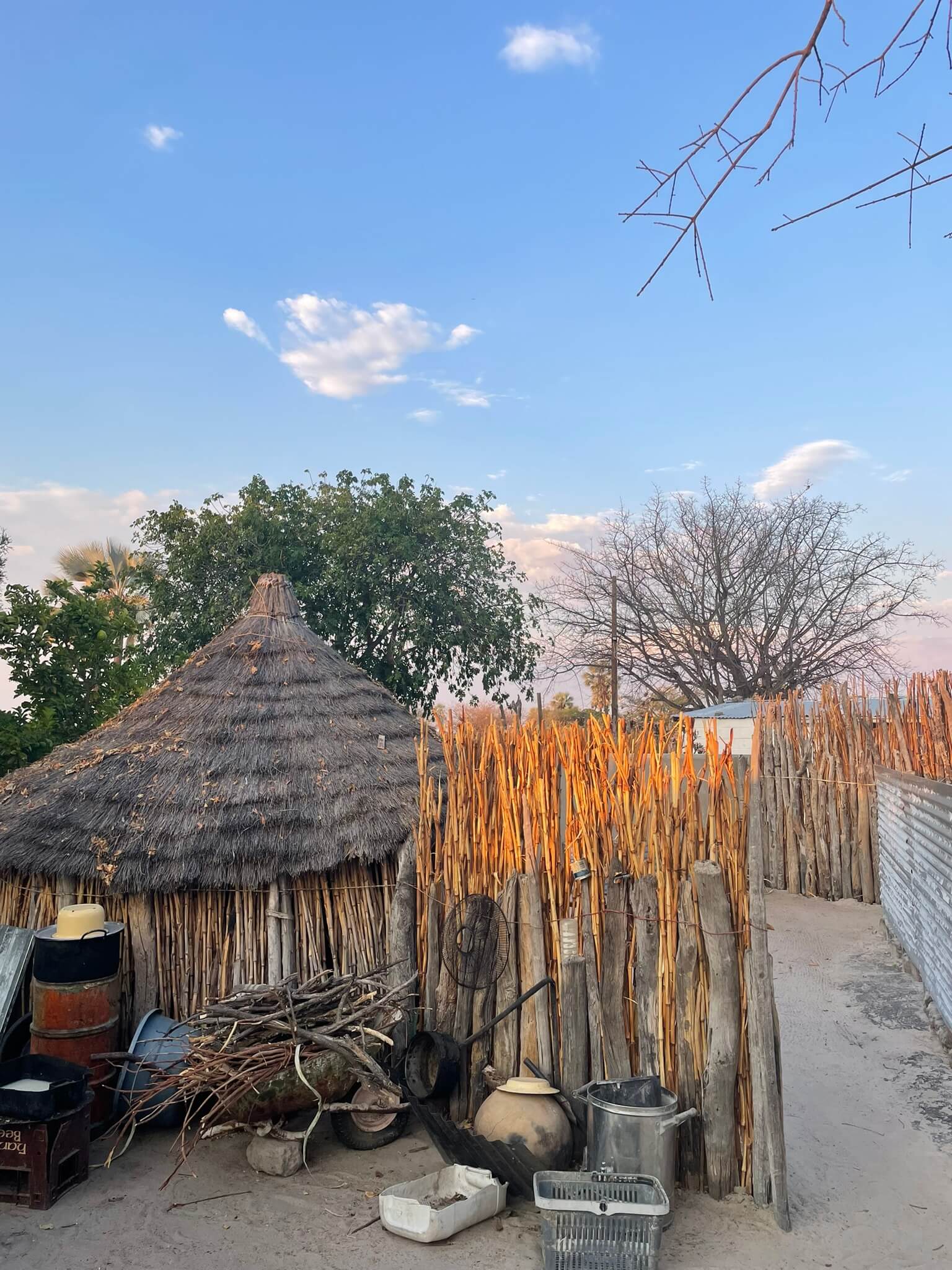 African thatched-roof hut behind a tall stick fence with scattered containers and firewood in the sandy yard